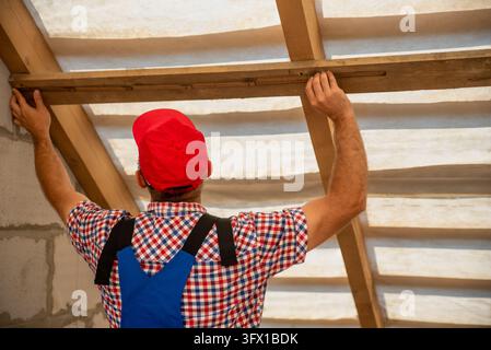 Construction worker in a red cap and work uniform carefully measures and installs a wooden roof beam inside a new home, demonstrating skilled carpentr Stock Photo