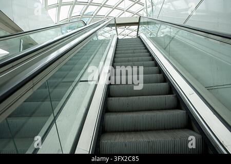 An escalator with transparent glass sides and steps, leading upward beneath a modern arched roof featuring a mesh-patterned glazed design. High qualit Stock Photo