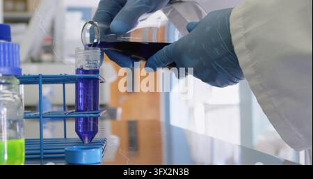 Pouring gloved hands lab coat transferring purple liquid from flask into conical tube at lab bench Stock Photo