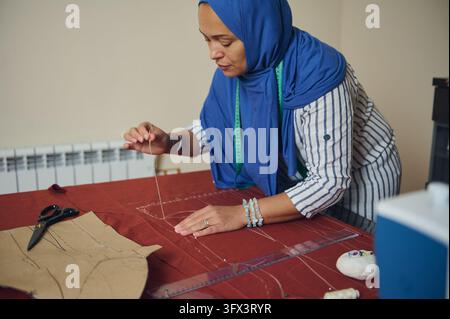 Woman tailor sews and measures red fabric for handmade clothing in a home tailoring workshop, using paper garment patterns, a needle, thread and measu Stock Photo