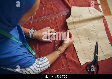 Woman tailor practices hand sewing on rust-red fabric, using a needle, thread, garment pattern and measuring tape at her home tailoring workshop for h Stock Photo