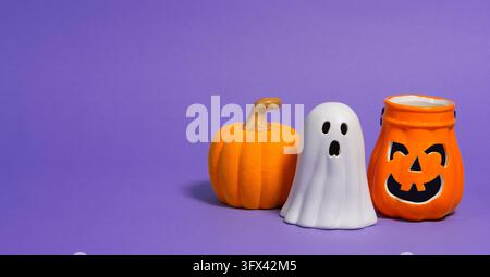 Front view of orange ceramic pumpkins with black faces, a small orange pumpkin, and a white ceramic ghost against a purple background Stock Photo