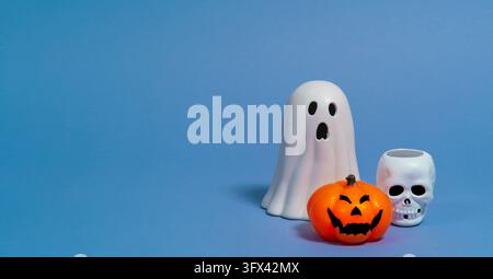 Front view of an orange ceramic pumpkin with a black face, a white skull, and a white ceramic ghost against a blue background Stock Photo