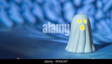 Front view of a white ceramic ghost with a yellow lit face set against a blue background with black shadows. Stock Photo