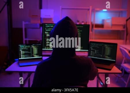 A mysterious hooded hacker working on a computer in a dark room. A concept for cybersecurity, hacking, cybercrime, and data privacy threats. Stock Photo