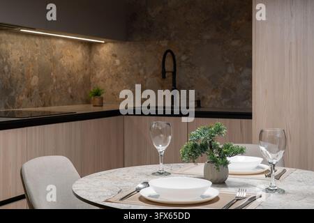 Modern dining area and kitchen interior featuring a round stone table set for two, wine glasses, and warm lighting Stock Photo
