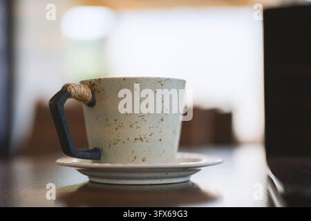A rustic ceramic coffee cup with a black metal handle wrapped in natural jute rope, resting on a matching saucer atop a wooden table in a softly lit café interior, shallow depth of field highlighting texture and craftsmanship Stock Photo