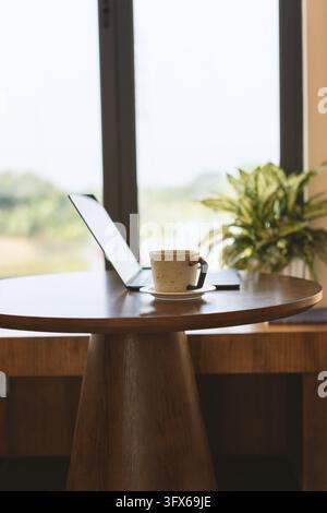 A minimalist modern workspace featuring an open laptop and a ceramic coffee cup on a round wooden table beside a large window with soft daylight, green plant in background, evoking calm productivity and remote work ambiance in a contemporary urban café or home office setting Stock Photo