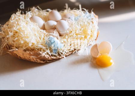 A rustic woven basket filled with shredded paper straw holding several white chicken eggs, one cracked open with bright yellow yolk spilling onto a light surface, accompanied by delicate blue floral decorations—evoking an Easter celebration or springtime food preparation scene in a natural, softly lit kitchen setting Stock Photo