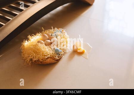 A decorative Easter nest made of woven straw and ceramic, filled with pastel-colored eggs and small blue ornaments, placed on a smooth beige countertop beside a cracked egg with spilled yolk and white, illuminated by soft natural light from a nearby window with horizontal blinds casting diagonal shadows. Stock Photo
