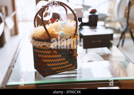 A handwoven decorative gift basket with zigzag pattern in brown, yellow, and blue, filled with shredded paper and small pastel eggs, adorned with white lace trim, a yellow fabric flower, and a sheer ribbon bow, placed on a modern glass-top coffee table in a bright indoor setting Stock Photo