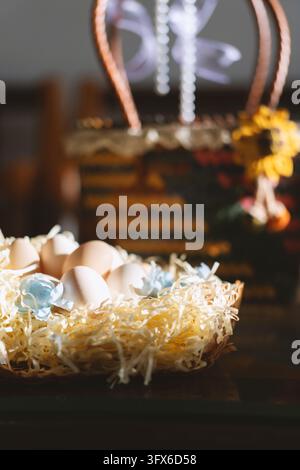A close-up of natural brown and white Easter eggs nestled in a woven wicker basket lined with pale yellow shredded paper straw, accented with delicate blue floral ribbons, set on a dark wooden table with soft backlighting and a blurred festive background featuring a decorative Easter basket and yellow flowers, evoking a warm, traditional spring holiday atmosphere in a home setting. Stock Photo