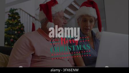 Sitting couple using laptop at home wearing Santa-hats pink-polo tree Merry Christmas overlay Stock Photo