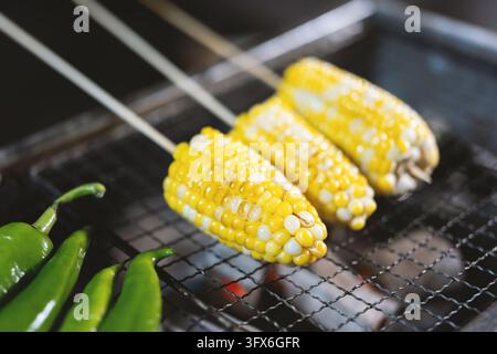 Three ears of sweet corn on wooden skewers grilling over glowing charcoal on a metal wire rack, accompanied by fresh green chilies resting beside the grill, evoking a vibrant summer street food scene with warm lighting and appetizing char marks Stock Photo