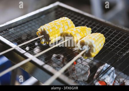 Three ears of sweet corn on wooden skewers grilling over glowing charcoal briquettes on a metal mesh barbecue grate, vibrant yellow kernels with slight charring, shallow depth of field highlighting texture and steam, typical of Southeast Asian street food culture in Thailand or Vietnam Stock Photo