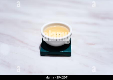 A small white ceramic teacup filled with pale golden oolong tea, resting on a dark green square ceramic saucer, placed on a light veined marble surface in soft diffused lighting, evoking tranquility and traditional East Asian tea culture Stock Photo