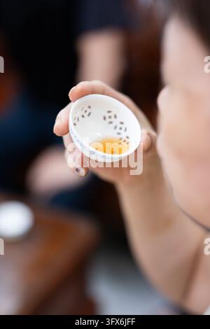 A person holding a small white perforated ceramic tea cup filled with amber-colored tea during a traditional East Asian tea tasting session, close-up focus on the cup and fingers, shallow depth of field with blurred background suggesting a quiet indoor setting Stock Photo