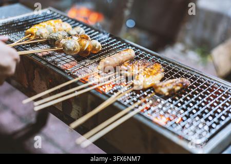 Grilled yakitori skewers including chicken thigh, sausage, shiitake mushrooms, and quail eggs sizzling over glowing charcoal on a traditional Japanese metal grill at a night market stall in Tokyo, with a vendor’s hand adjusting the skewers under warm ambient lighting Stock Photo