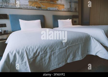 Two neatly made twin beds with white linens and blue upholstered headboards in a contemporary hotel room, featuring a large wall-mounted cityscape artwork above the beds and warm ambient lighting, Shanghai, China Stock Photo