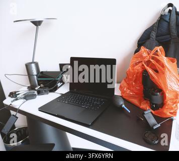 Modern minimalist workspace with a black laptop on a white-and-black desk, accompanied by photography gear including two camera lenses in an orange plastic bag, a blower bulb, lens cap, USB hub, power adapter, and a sleek metallic desk lamp; background features a dark fabric tote bag and clean white wall, suggesting a portable creative setup in an urban apartment or hotel room Stock Photo