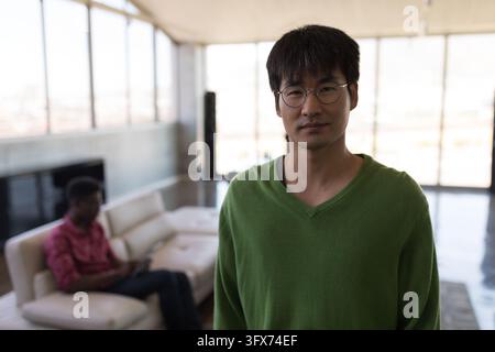 Portrait of casually dressed Asian male executive standing in office with colleague working in the background Stock Photo