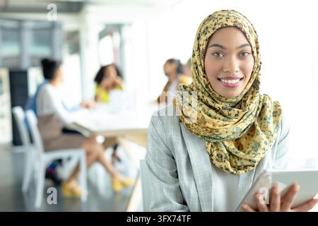 A young woman in hijab is holding a tablet, smiling at camera. Her friends are discussing in the background, in a bright office space, unaltered Stock Photo