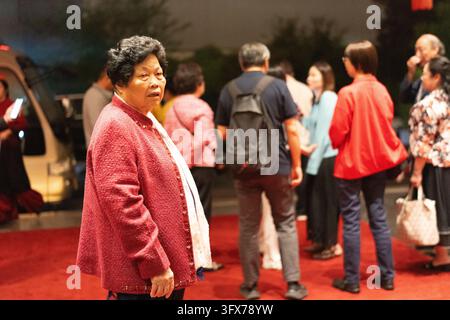 An elderly East Asian woman in a textured red jacket stands on a red carpet at an outdoor evening event, surrounded by a casually dressed crowd with soft ambient lighting and a vehicle visible in the background Stock Photo