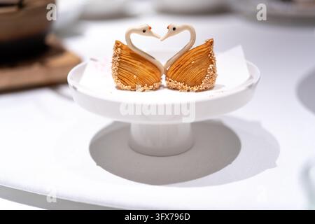 Two intricately crafted swan-shaped pastries made of flaky layered dough and decorated with sesame seeds, their necks curved to form a heart shape, presented on a white ceramic pedestal dish in a high-end Chinese restaurant setting Stock Photo