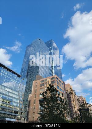Vertical view of a blue glass skyscraper rising behind brick buildings and an evergreen tree under white clouds, Manhattan, New York City. Stock Photo