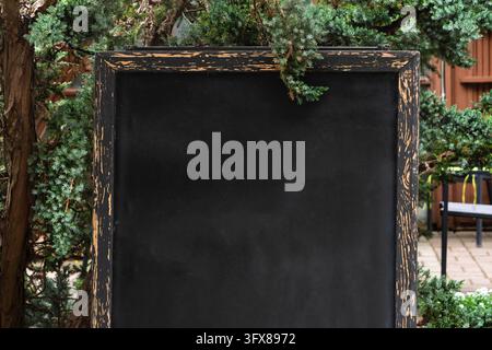 Weathered empty chalkboard with distressed wooden frame stands in lush green garden, inviting creativity and outdoor announcements. Stock Photo