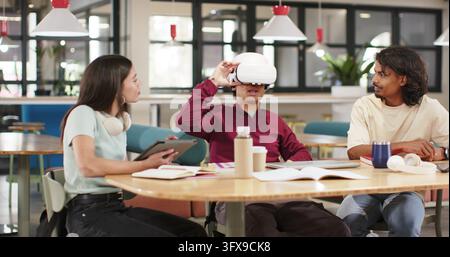 African American peers sitting around wooden table in study hub, center man using white VR headset. Coworking, collaboration, virtual, goggles, lounge Stock Photo