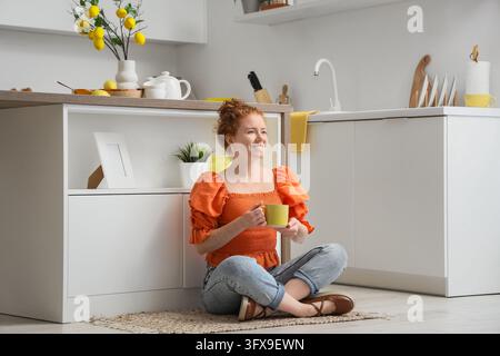 Mindfulness and relaxation at home, happy young woman sitting on kitchen floor with a cup of coffee enjoying a peaceful morning in modern interior Stock Photo