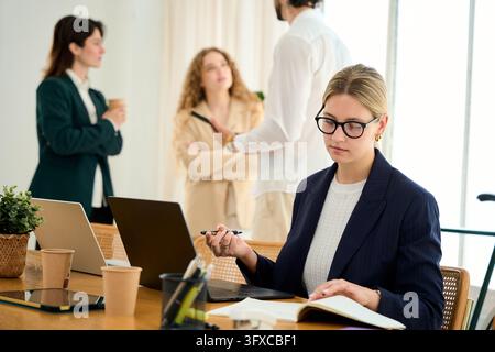 Focused businesswoman writing in notebook, holding pen while working on laptop. Colleagues collaborating in background Stock Photo