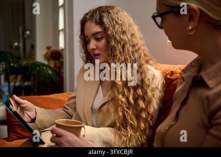 Two businesswomen discussing project strategy, looking at a digital tablet, sitting on a sofa during an informal meeting Stock Photo