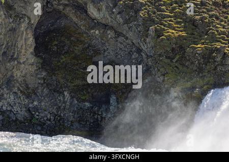 River spray rises beside a fractured, moss-covered rock wall as white water rushes through the Godafoss area on a sunny day in North Iceland. Stock Photo