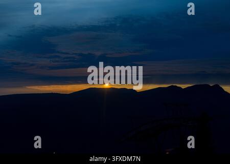 Vancouver, Canada - Horizontal panoramic capture of bright orange sun setting behind layered mountain silhouettes beneath deep blue nightfall skies. Stock Photo