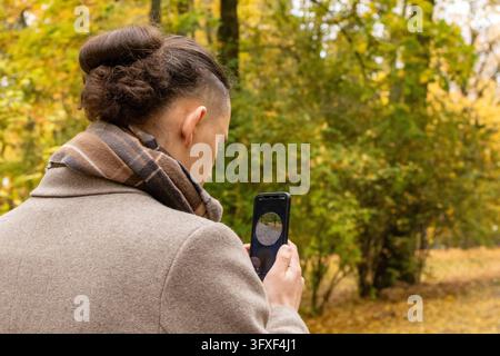 Man with a hair bun in a beige coat records a video message on his smartphone while standing on an autumn park path. Concept of staying connected and Stock Photo