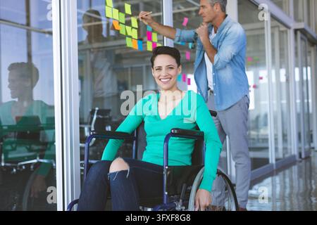 Middle-aged woman in wheelchair smiling, man placing sticky notes. She has short black hair, he wearing casual shirt and jeans, unaltered Stock Photo