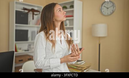 Woman holds tablet with hands and looks up in an office building while checking documents on desk; thoughtful reflection. Stock Photo
