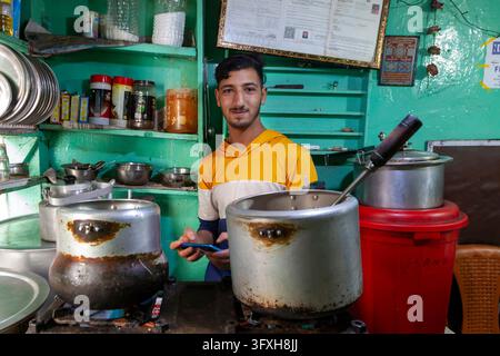 Young man cooking with large pots in a small green kitchen and holding a phone, Ladakh, Leh, India Stock Photo