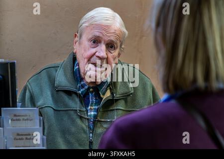 An elderly man with white hair is speaking at a bar in Castelo Rodrigo, in the district of Guarda, Portugal. The woman he is talking to, the journalist Angela Berg, can be seen from behind. On the left are leaflet holders bearing the inscription ‘Bienvenido a Castelo Rodrigo’ (in English: Welcome to Castelo Rodrigo). Stock Photo