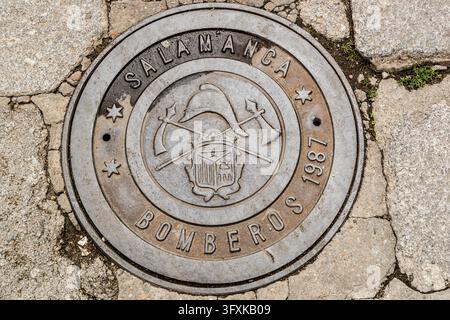 A metal cover inscribed “SALAMANCA BOMBEROS 1987” (In English: fire brigade 1987) lies in the stone paving of Salamanca in Castile and León, Spain. In the centre it shows a firefighter’s helmet above two crossed axes and a heraldic shield. Three stars stand on the ring. Stock Photo