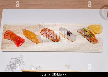 Five pieces of nigiri sushi featuring various raw fish toppings arranged on a textured rectangular plate with pickled ginger. Stock Photo