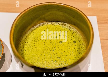 A close-up, top-down view of a ceramic bowl filled with frothy green matcha tea resting on a white napkin. Stock Photo