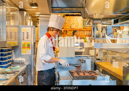 Tokyo, Japan, NOV 22, 2024 - A chef wearing a tall white hat and red neckerchief uses tongs to cook meat patties on a grill in a busy restaurant kitch Stock Photo