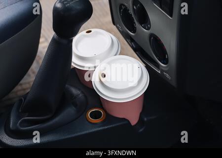 Two takeaway coffee cups with white plastic lids in car cup holders beside gear lever. Hot drinks inside vehicle during road trip and daily commute. Stock Photo
