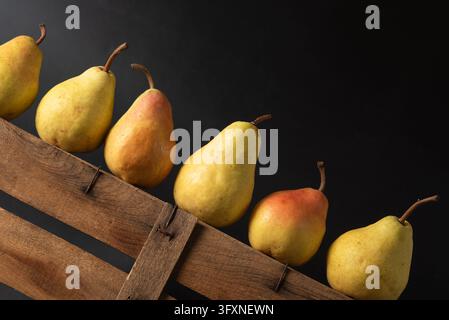 A variety of fresh pears in different shades of yellow and orange are neatly arranged on a wooden crate. The dark background enhances the vibrant colo Stock Photo