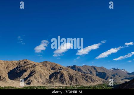 A wide landscape shot showcasing a line of small white clouds floating above towering brown mountain ranges in an arid desert valley of Ladakh. Stock Photo