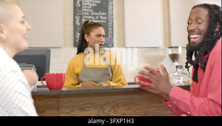 Diverse barista and customers standing at coffee shop counter, barista wearing gray apron, red mugs. Cafe, interior, brick, chalkboard, espresso, grin Stock Photo