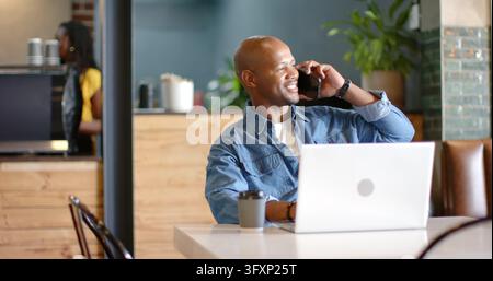 Talking man wearing denim shirt holding smartphone at cafe table, using laptop with takeaway cup. Coffeehouse, phone, computer, plant, wood, warmth, m Stock Photo